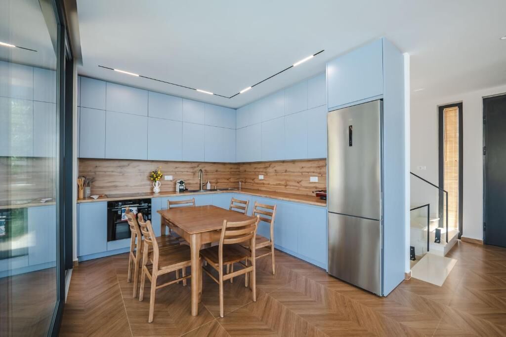 A modern kitchen with blue cabinets, a wooden dining table, and a stainless steel refrigerator.