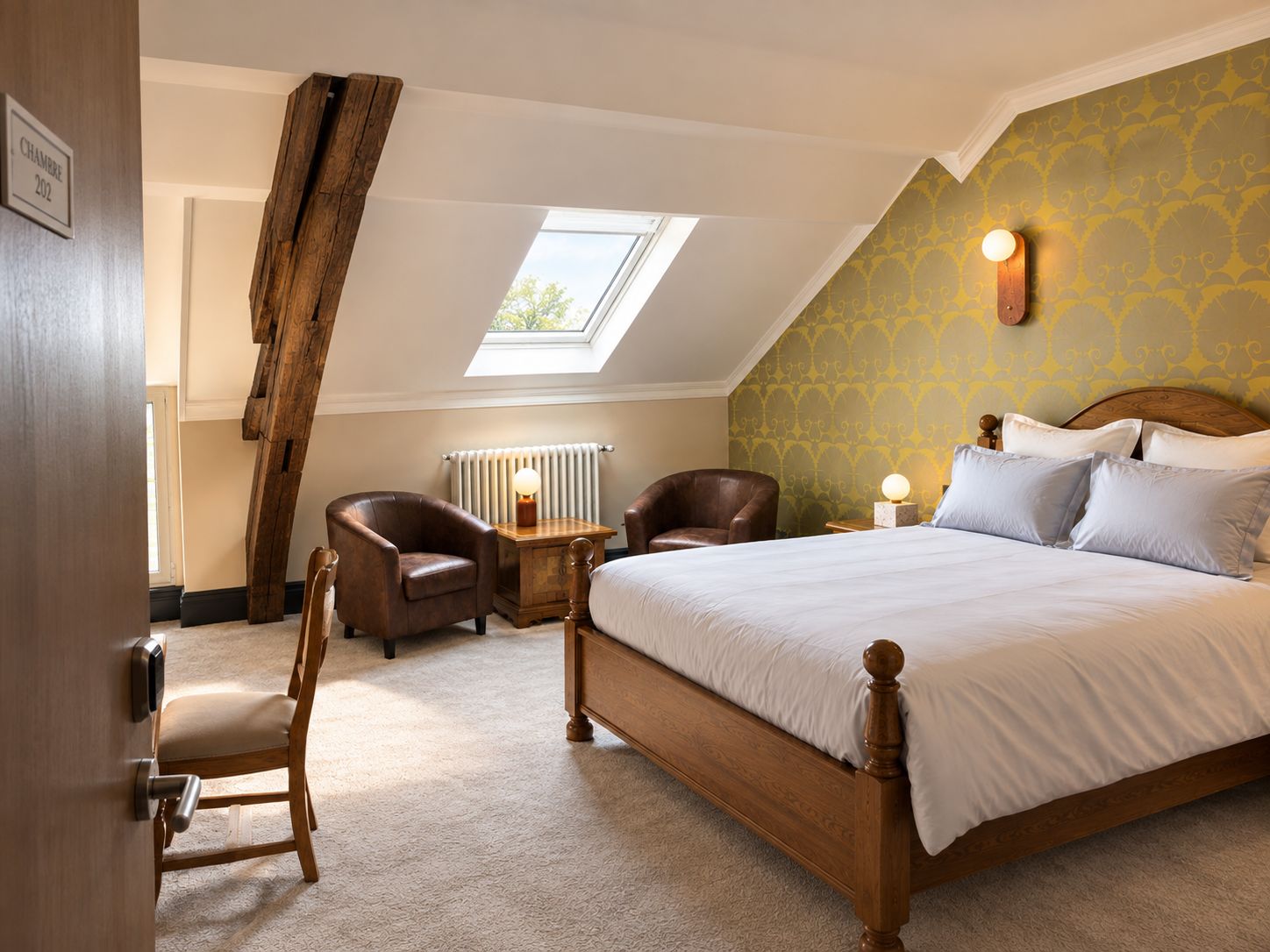 A bedroom with a sloped ceiling, a skylight, a bed with white bedding, two leather chairs, a wooden nightstand with a lamp, and a radiator.
