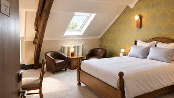 A bedroom with a sloped ceiling, a skylight, a bed with white bedding, two leather chairs, a wooden nightstand with a lamp, and a radiator.