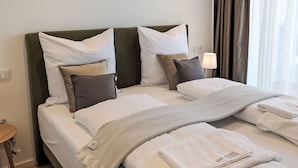 Ein ordentlich bezogenes Bett mit weißen Bettwäsche und einer grauen Tagesdecke, ein Nachttisch mit Lampe sowie Blick durch Vorhänge aus einem Fenster.