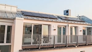 Ein modernes Gebäude mit großen Fenstern, einem Balkon mit Metallgeländern und Solaranlagen auf dem Dach.