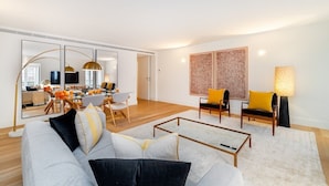 Un salon moderne avec un canapé, une table basse et des fauteuils. On y trouve également un coin repas avec une table et des chaises, et une cuisine à l’arrière-plan.