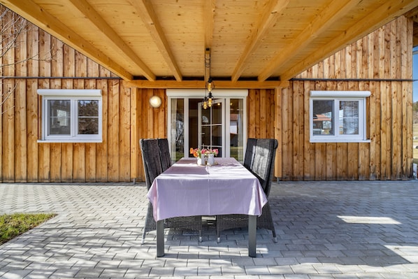 A wooden exterior building with a covered patio area featuring a table with chairs and a vase of flowers.