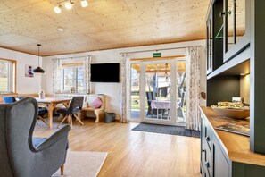 A room with a wooden ceiling, a dining area with a table and chairs, a television mounted on the wall, and a kitchen area with a counter and cabinets.