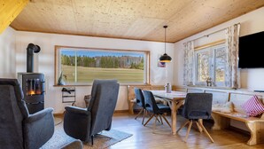Tunnelmallinen olohuone, jossa on takka, mukavat istuimet, ruokailutila ja suuri ikkuna, josta avautuu näkymä metsäiselle alueelle.