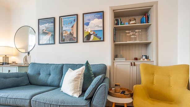 Una sala de estar con un sofá azul, un sillón amarillo, una estantería y tres carteles enmarcados en la pared.