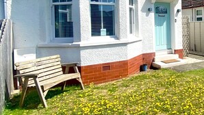 Una casa blanca de dos pisos con una puerta azul, un pequeño porche con un banco y un jardín con flores amarillas.