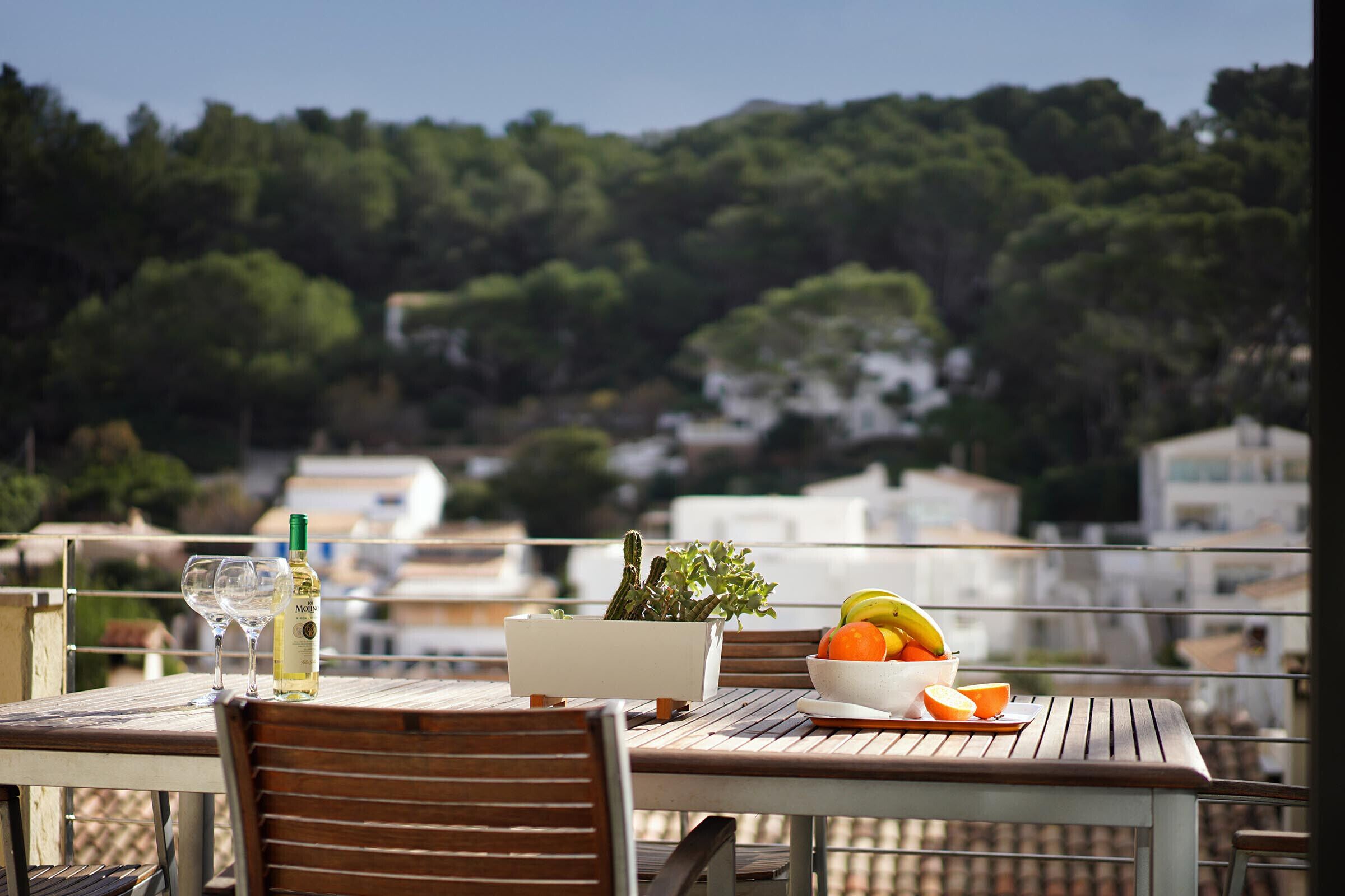 A table with a bottle, a glass, a bowl of fruit, and a potted plant on a balcony overlooking a residential area.