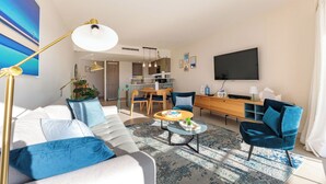 Uma sala de estar moderna com sofá, poltronas, mesa de centro e televisão. Há uma área de jantar com mesa e cadeiras, e uma cozinha ao fundo.