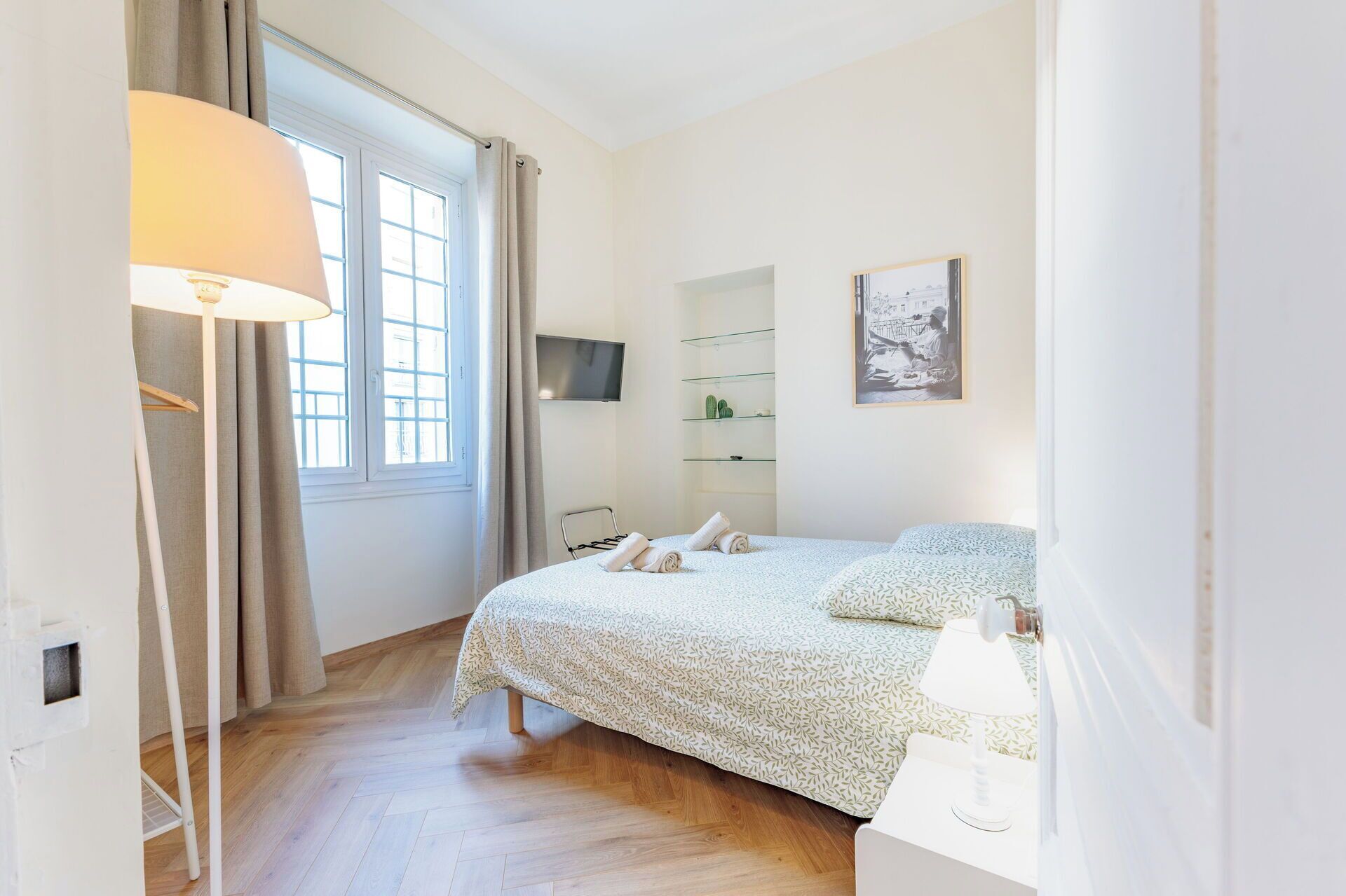 A bedroom with a bed, a bedside table, a lamp, a TV, and a window with curtains.
