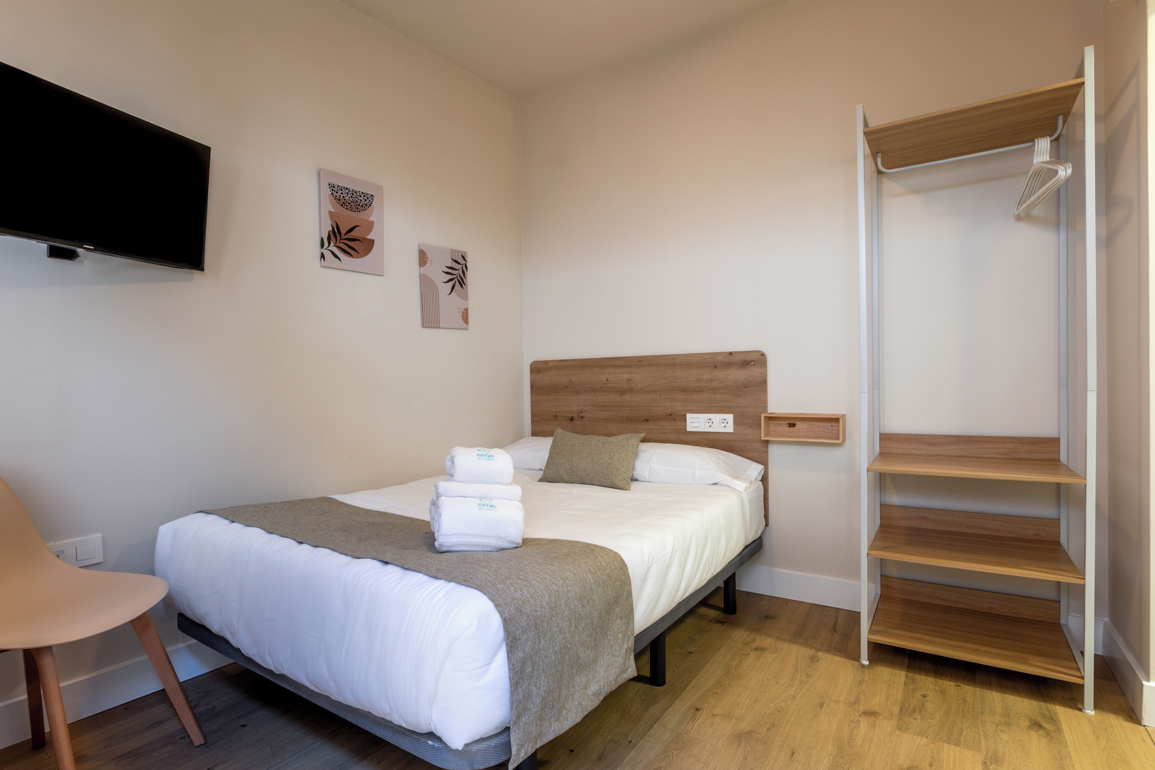Une chambre d’hôtel bien rangée, avec un lit, une chaise, un téléviseur et une étagère en bois.