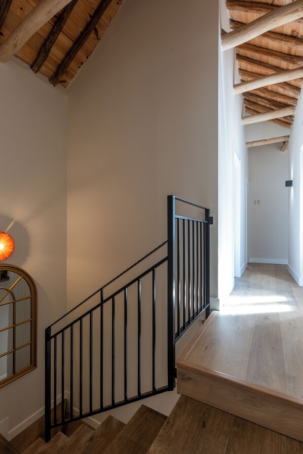 Een trap met houten treden en een zwarte metalen balustrade, leidend naar een kamer met een houten plafond en een lamp aan de muur.