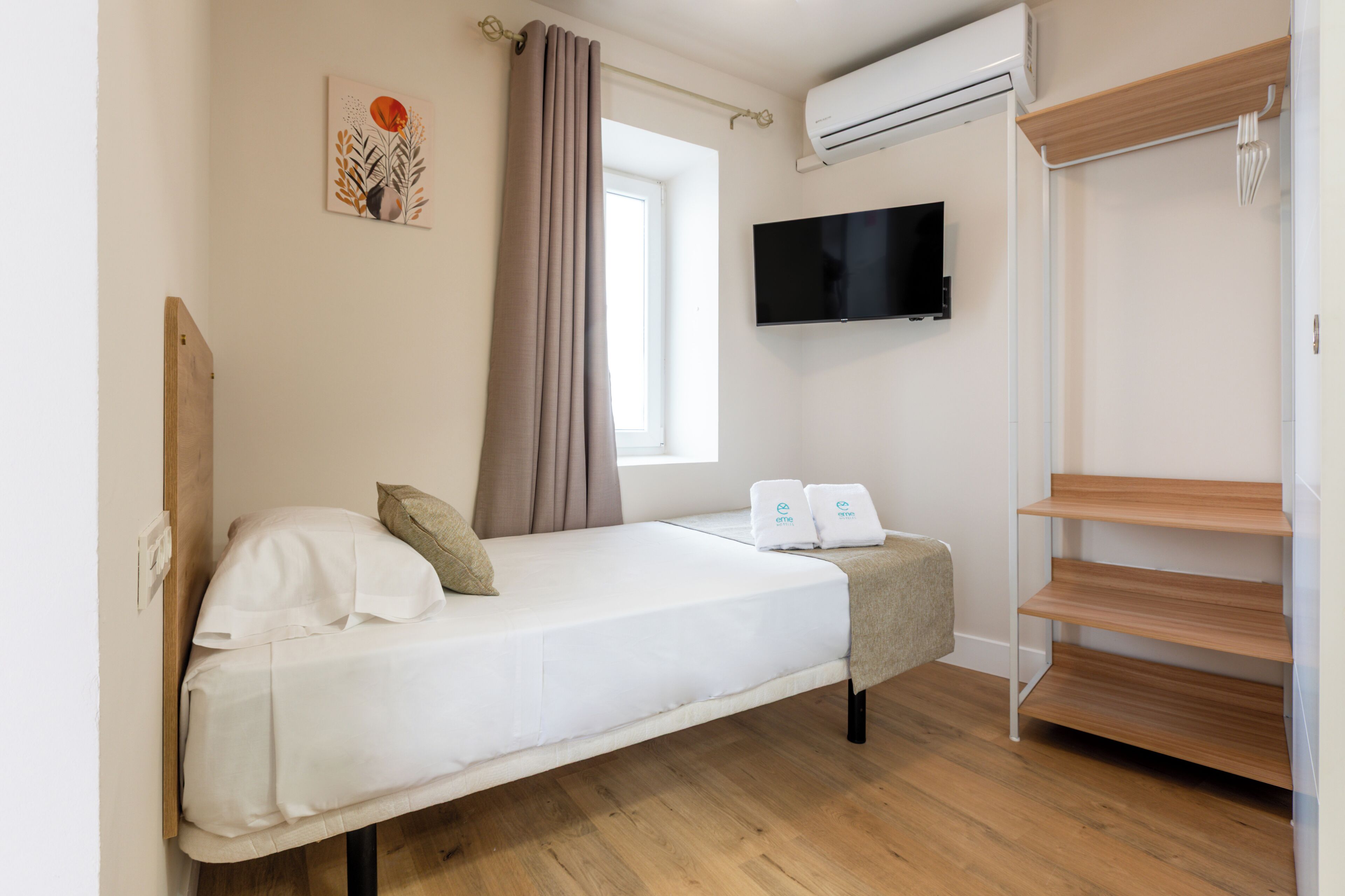A single bed with a TV, an air conditioner, and a window with curtains.