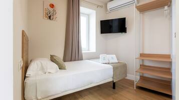 A single bed with a TV, an air conditioner, and a window with curtains.