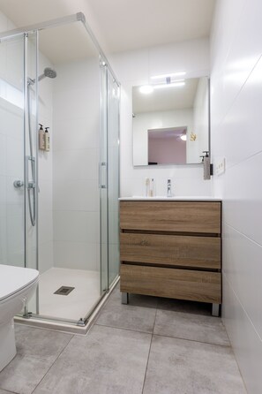 Un baño con cabina de ducha de vidrio, inodoro blanco, tocador de madera con cajones y espejo sobre el tocador.