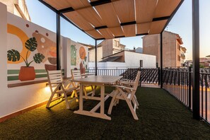 A rooftop terrace with a wooden table and chairs, a potted plant, and a mural on the wall. - Hospedería Boho Granada by eme hoteles (La Zubia)