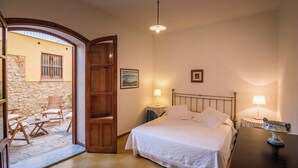 A bedroom with a bed, two bedside lamps, a wooden door, a stone wall, and a small table with chairs outside.