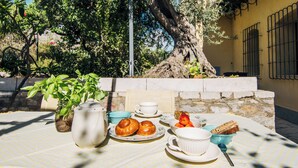 A table set with a teapot, cups, plates, and food items, with a large tree and stone wall in the background.