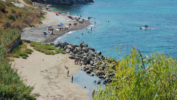 A beach with people swimming and sunbathing, surrounded by rocks and greenery.