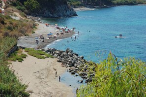 A beach with people sunbathing and swimming, rocky shoreline, and a clear blue sea.
