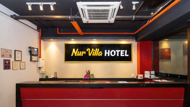 A hotel reception desk with a red counter, a large illuminated sign reading 'Nur Villa Hotel,' and framed certificates on the wall. - Capital O 91277 Nur Villa Hotel (Malacca City)