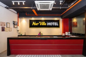 Eine Hotelrezeption mit einer roten Theke, einem großen, beleuchteten Schild mit der Aufschrift 'Nur Villa Hotel' und gerahmten Zertifikaten an der Wand.