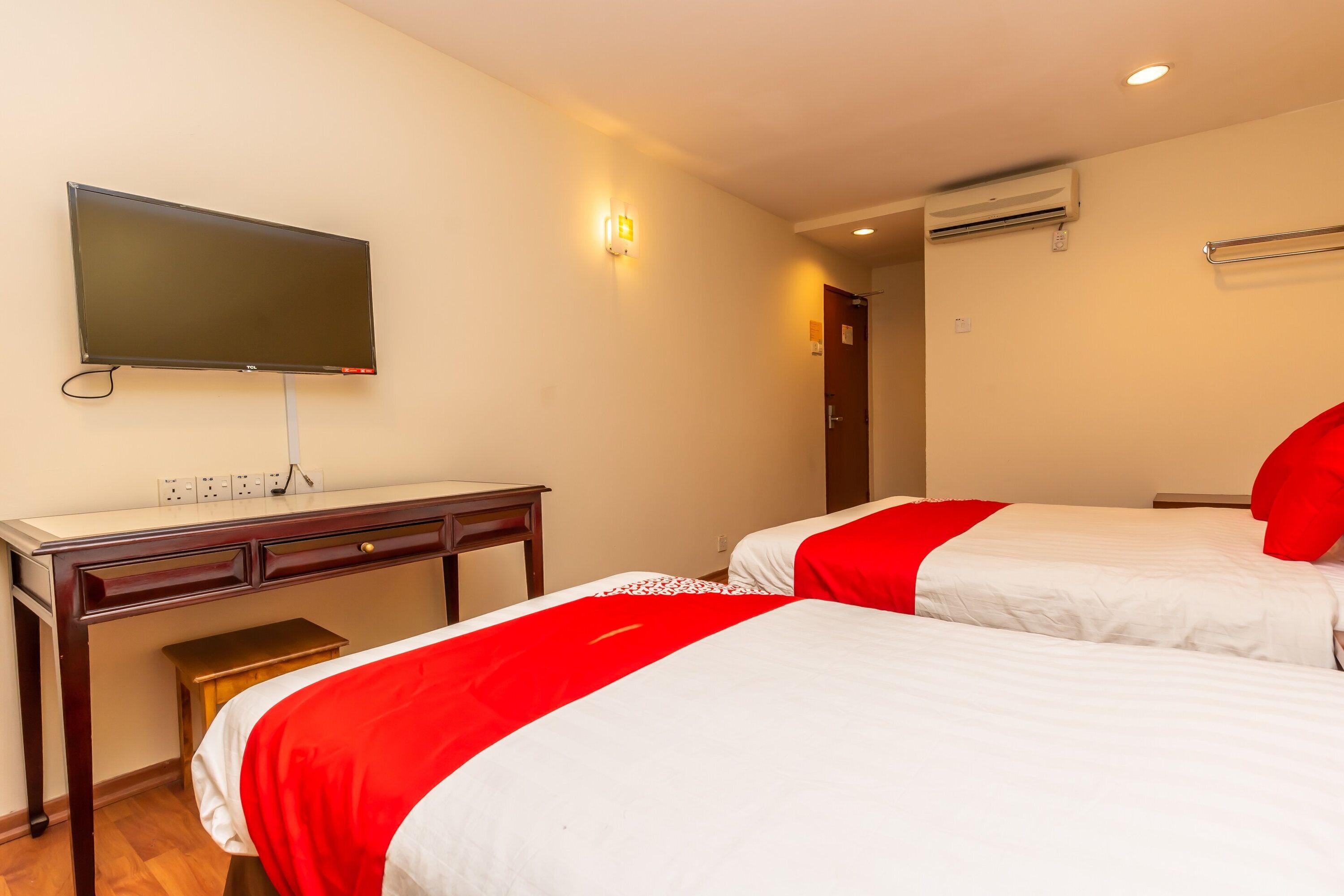 Une chambre d’hôtel avec un téléviseur fixé au mur, un bureau avec des tiroirs, deux lits avec une literie rouge et blanche, et un climatiseur fixé au mur.