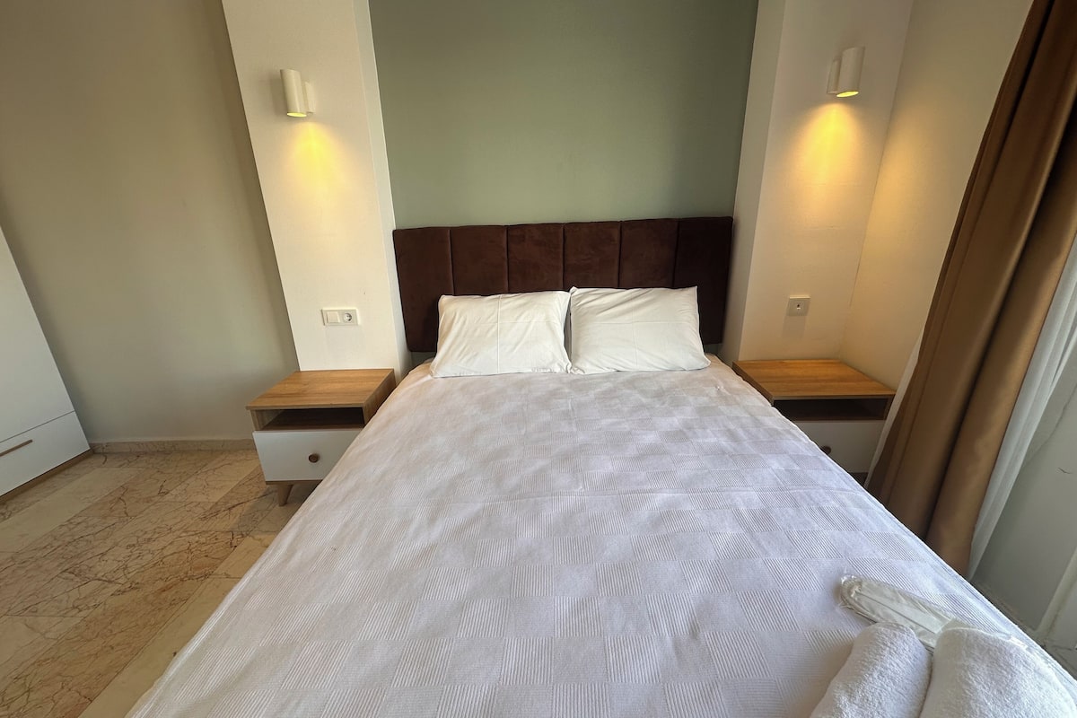 A neatly made bed with a headboard, two bedside tables, and a nightstand with a drawer.