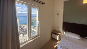 Ein Schlafzimmer mit Bett, Nachttisch, Fenster mit Blick auf Meer und Berge sowie einem Vorhang.