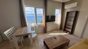 Ein Zimmer mit einem Holztisch, weißen Stühlen, einem gelben Sofa, einem Fernseher an der Wand und Meerblick durch große Fenster.