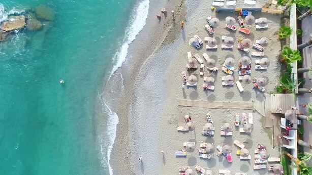 An aerial view of a beach with rows of sun loungers and umbrellas, a rocky shoreline, and a few people in the water. - Club Monte Carlo Hotel - All Inclusive (Alanya)