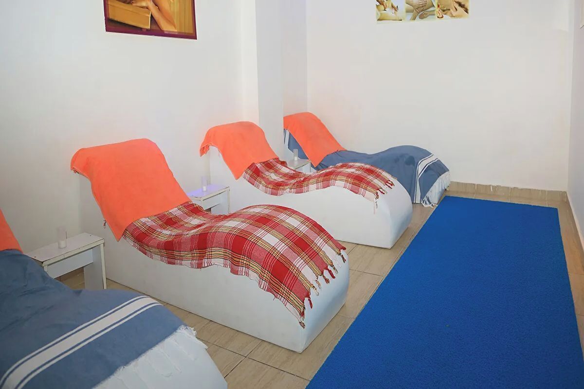 A room with three beds, each with a different colored headboard. There is a small table with a lamp next to the first bed.