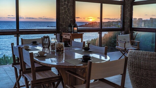 A dining area with a table set for two, overlooking a sunset view of the sea.