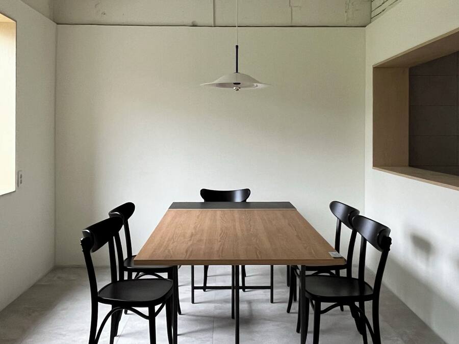 A conference room with a wooden table, four black chairs, a hanging light fixture, and a window on the left.