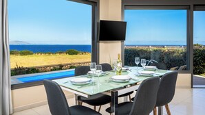 A dining area with a table set for four, large windows with a view of the sea, and a flat-screen TV mounted on the wall. - Aqua Natura Villas - Villa Aqua (Rhodes)
