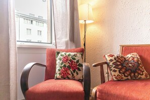 Une pièce avec deux fauteuils rouges, dont l’un est agrémenté d’un coussin à motifs floraux et l’autre d’un coussin à motifs géométriques. Un lampadaire est placé entre les fauteuils, et une fenêtre avec des rideaux est visible à l’arrière-plan.