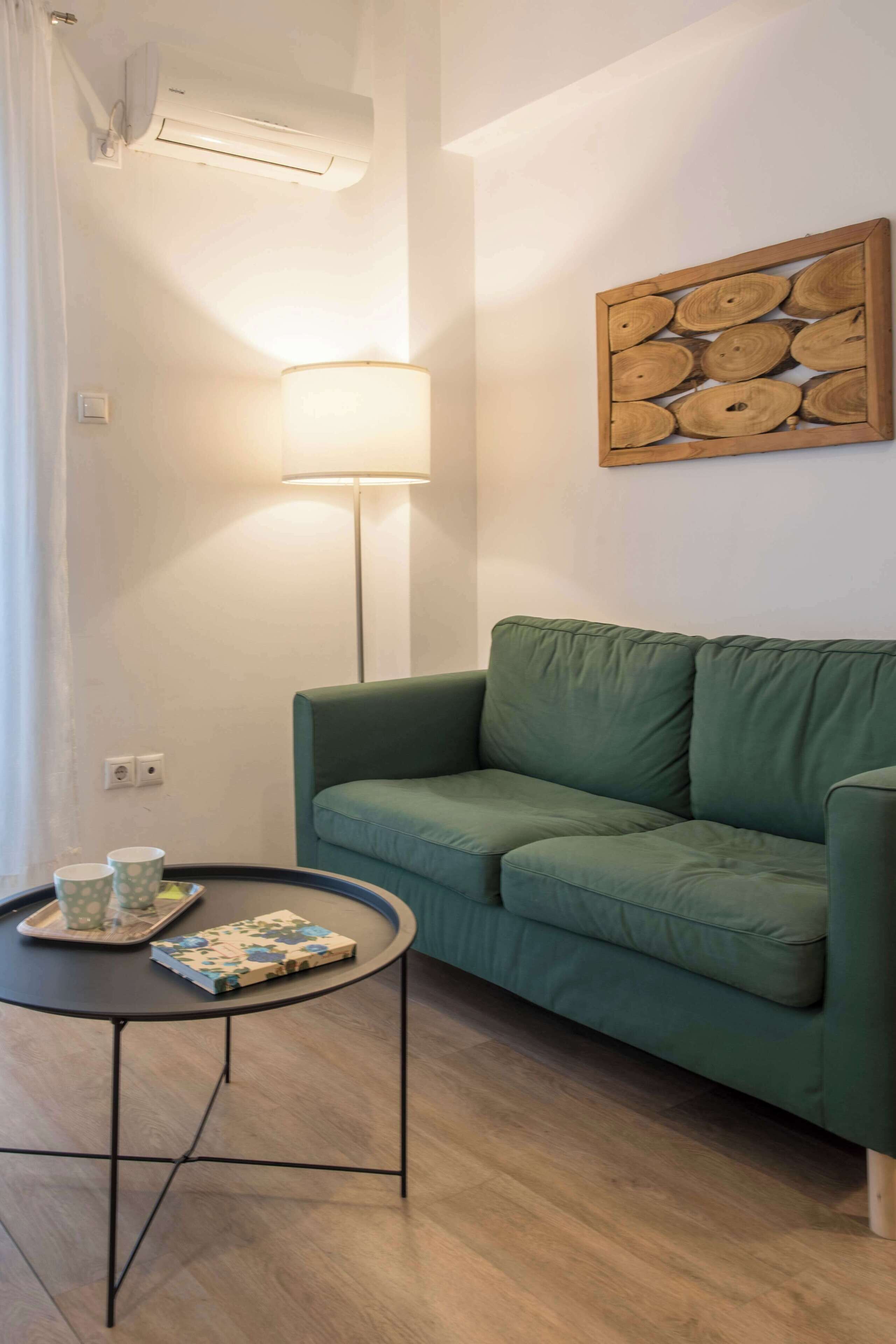 Ein grünes Sofa, ein kleiner runder Tisch mit Tablett und Buch, eine Stehlampe und ein hölzernes Wandbild.