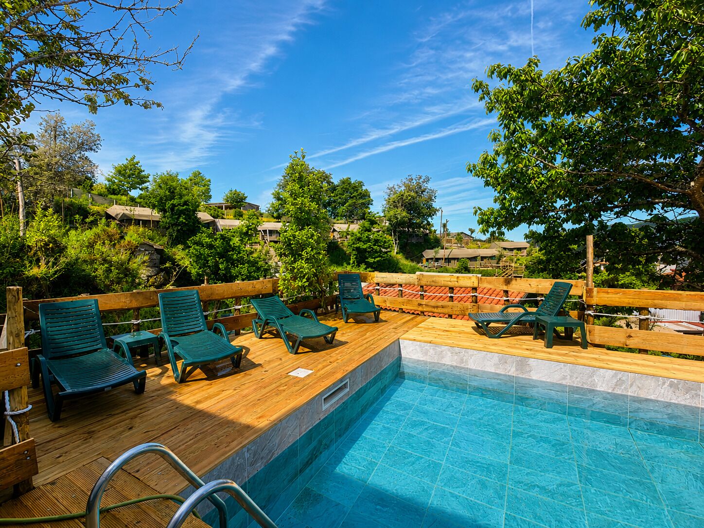 A poolside area with wooden deck, green lounge chairs, and a clear view of the surrounding greenery and buildings.