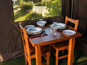 A wooden dining table set for four with plates, glasses, and cutlery, positioned in a tent-like structure with a view of greenery outside.
