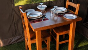 Une table de salle à manger en bois, dressée pour quatre personnes avec des assiettes, des verres et des couverts, placée à l’intérieur d’une structure ressemblant à une tente, d’où l’on peut admirer la verdure environnante.