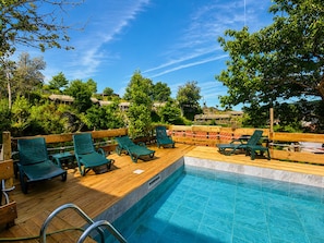 Un espace aménagé au bord de la piscine, avec une terrasse en bois, des chaises longues vertes et une vue dégagée sur la végétation et les bùtiments environnants.