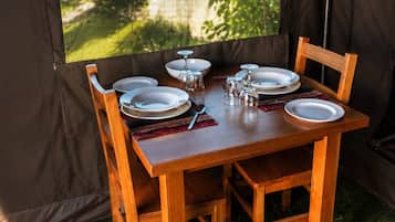 A wooden dining table set for four with plates, glasses, and cutlery, positioned in a tent-like structure with a view of greenery outside.