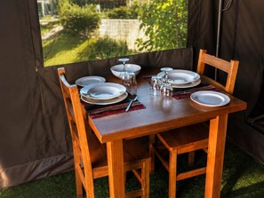 Une table de salle Ă manger en bois, dressĂ©e pour quatre personnes avec des assiettes, des verres et des couverts, placĂ©e Ă lâintĂ©rieur dâune structure ressemblant Ă une tente, dâoĂč lâon peut admirer la verdure environnante.