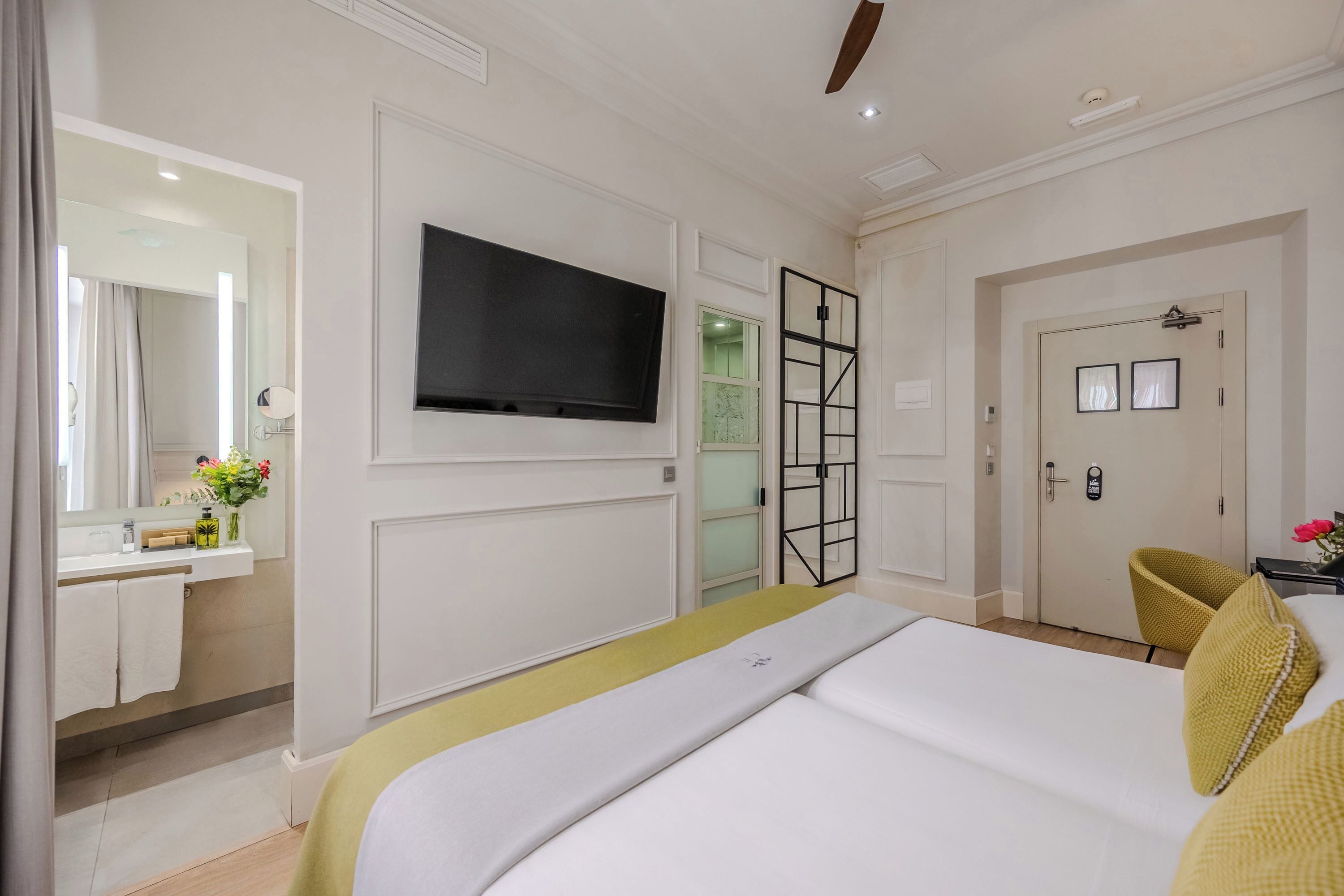 Ein Hotelzimmer mit Bett, Wand-Fernseher, Badezimmer mit Spiegel und Waschbecken sowie einer Tür mit Griff.