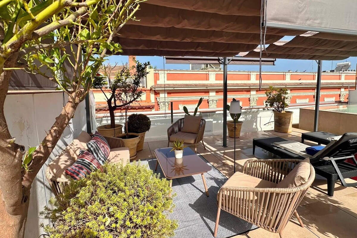 A rooftop terrace with outdoor furniture, a potted plant, and a view of buildings in the background.