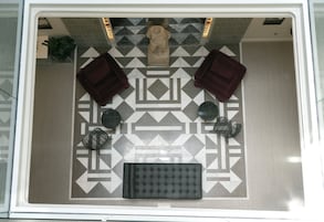 Vista cenital de un vestíbulo de hotel con piso estampado, un área de descanso central con un banco acolchado, dos sillones y una pequeña estatua.