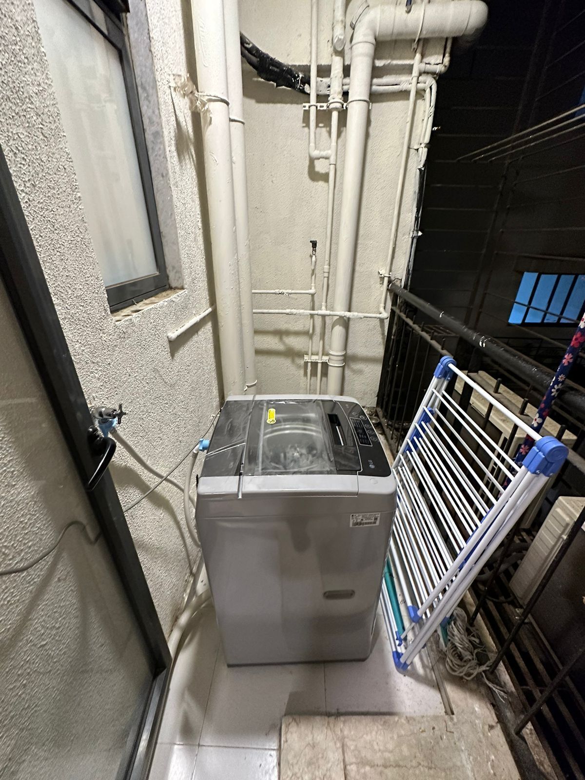 A compact, industrial-style laundry room with a large, grey washing machine, visible pipes, and a metal rack.