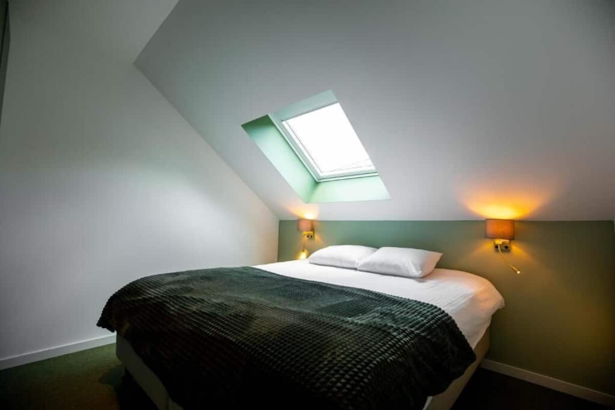 Une chambre avec un puits de lumière, un lit recouvert d’une couverture sombre, deux lampes de chevet et un mur d’accent vert.