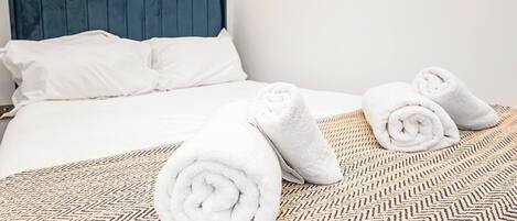 A neatly made bed with white linens, a patterned throw, and rolled towels on a chevron-patterned bedspread.