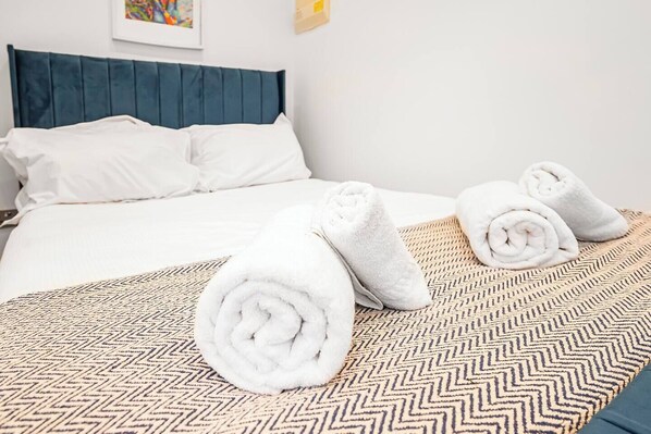 Una cama bien hecha con sábanas blancas, un plaid estampado y toallas enrolladas sobre una colcha con estampado en zigzag.